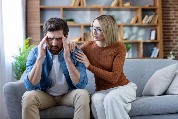 Bir kadın nazikçe erkeğin omzuna dokunuyor, ona destek oluyor ve baş ağrısı çekiyor ve modern bir oturma odasındaki kanepede stresli hissediyor.