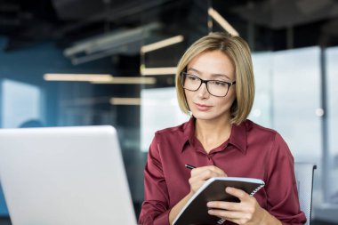 Kendine güvenen iş kadını, dizüstü bilgisayara bakarken gözlük takıyor ve modern ofis ortamında çalışmaya ve planlamaya odaklanıyor.