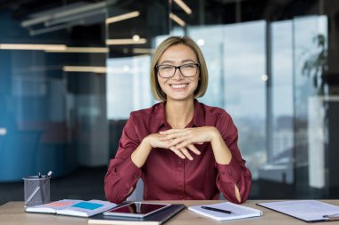 Başarılı genç profesyonel kadın ofis masasında oturuyor, gözlük takarken izleyiciye güvenle gülümsüyor, kurumsal başarıyı ve modern çalışma alanındaki olumlu tutumu simgeliyor.
