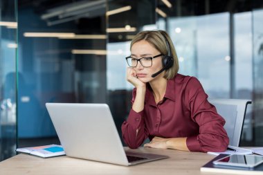Kadın müşteri hizmetleri görevlisi kulaklık ve gözlük takıyor. Ofis masasında dizüstü bilgisayarla çalışırken yorgun ve sıkılmış görünüyor. Stres, yorgunluk ve tükenmişlik taşıyor.