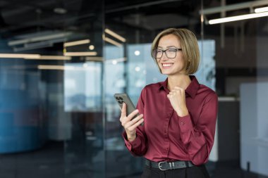 Gözlüklü profesyonel iş kadını, akıllı telefonuna bakarken heyecanlanıyor ve başarısını kutluyor. Çağdaş ofis ortamında bir iş anlaşması kazanıyor.