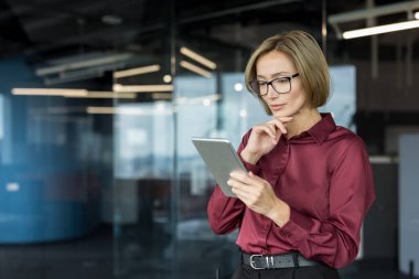 İş kadını gözlüklü ve bordo gömlekli bir kadın çağdaş bir ofiste dikiliyor, dijital tablet üzerindeki verileri dikkatle inceliyor odaklanmış ve derin düşüncelere dalmış bir ifadeyle