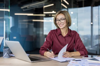 Profesyonel iş kadını gülümsüyor, gözlük takıyor ve masasındaki belgelere bilgisayar ve çağdaş ofis ortamında çeşitli evraklarla not alıyor.