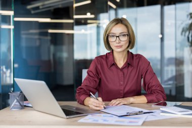 Modern kurumsal ofisteki kendine güvenen Asyalı iş kadını bilgisayarında ve gözlükleriyle belgeleri inceliyor, finansal planlama ve sözleşme imzalamaya odaklanmış ve hazır.