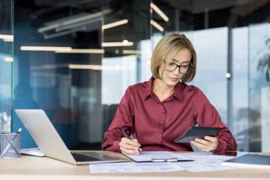 Resmi aşınma ve gözlüklü kadın muhasebeci hesap makinesi kullanarak finans istatistiklerini dikkatlice hesaplıyor ve ofis masasındaki belgelere dizüstü bilgisayar ve çeşitli kağıtlarla yazı yazıyor.
