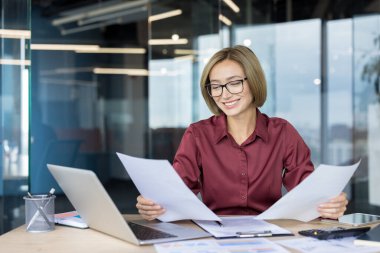 Modern bir ofis masasında gözlüklü deneyimli bir iş kadını finansal belgeleri ve raporları dizüstü bilgisayar ve hesap makinesiyle incelerken gülümsüyor.