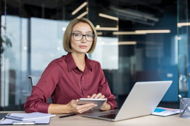 Kendine güveni tam bir iş kadını dijital tabletinde çalışırken, elinde bir dizüstü bilgisayarla, şık ofis ortamında profesyonellik ve modern teknoloji kullanımı sergiliyor.