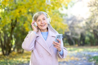 Yaşlı bir kadın, kablosuz kulaklıkla müzik dinlerken gülümsüyor, bir sonbahar parkında sarı yapraklı akıllı bir telefon tutuyor, huzurlu, neşeli, açık havada dinlenmenin keyfini çıkarıyor.