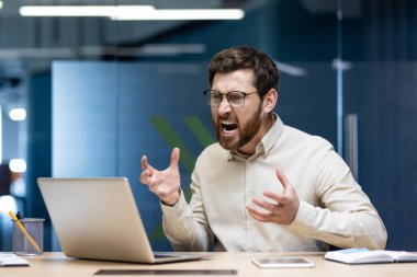 Öfkeli genç ofis çalışanı iş yerinde oturuyor ve dizüstü bilgisayara beceriksizce bağırıyor, elleriyle işaret ediyor..