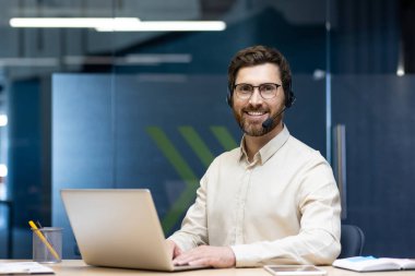 Ofisteki bir masada oturan, dizüstü bilgisayar kullanan ve kameraya güvenle bakan kulaklıklı genç bir adamın portresi..