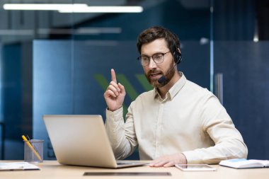 Ciddi bir genç adam, ofiste, kulaklıkla oturuyor ve bilgisayardaki bir video görüşmesinde konuşuyor, anlatıyor, işaret ediyor..