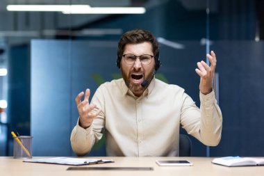 Ofiste oturan ve ellerini sallayarak kameraya bağıran öfkeli, kulaklıklı genç bir adam..