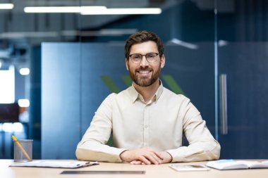 Hafif gömlekli ve gözlüklü gülümseyen genç bir iş adamının portresi ofiste bir masada oturmuş kameraya bakıyor..