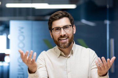 Ofiste oturan, kameraya konuşan ve el kol hareketi yapan genç bir adamın yakın plan portresi..