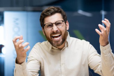 Gözlüklü ve gömlekli genç bir adamın yakın plan portresi. Ofiste, elleri ile kameraya işaret ederken öfkeyle konuşuyor ve bağırıyor..