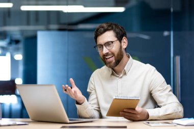 Ofiste çalışan gülümseyen genç bir adam, bir iş yerinde dizüstü bilgisayarın önünde oturuyor, elinde kalemle bir defter tutuyor, video toplantısı düzenliyor..