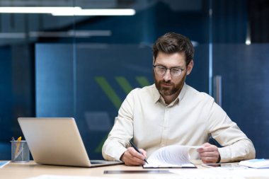 Odaklanmış ve ciddi bir genç adam ofiste çalışıyor, bir masada oturuyor ve belgeler ve kağıtlar yazıyor..