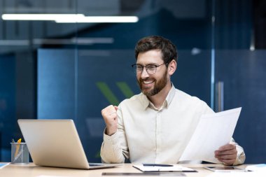 Mutlu genç bir işadamı ofiste bir masada oturuyor, elinde bir belge, dizüstü bilgisayara bakıyor ve eliyle zafer jesti yapıyor..
