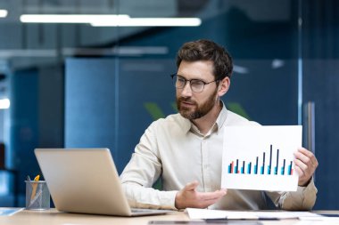 Ciddi bir genç adam bir ofiste çalışıyor, dizüstü bilgisayarın önünde bir masada oturuyor, video aracılığıyla konuşuyor, kameraya grafiklerle dokümanları gösteriyor ve sunuyor..