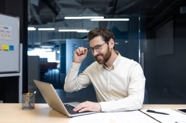 Sırıtkan genç ofis çalışanı masasında oturmuş elinde bardaklarla dizüstü bilgisayarın ekranına bakıyordu..