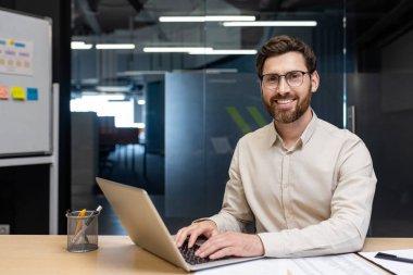 Gözlüklü ve açık renk gömlekli genç bir adamın portresi masada oturuyor, dizüstü bilgisayarla çalışıyor, gülümsüyor ve kameraya bakıyor..