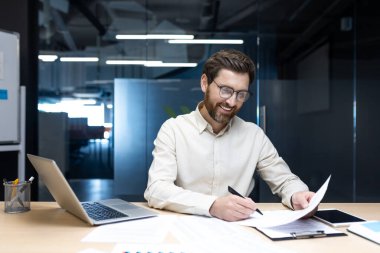 Başarılı ve gülümseyen genç bir işadamı ofiste elinde dizüstü bilgisayarla oturuyor ve belgeleri imzalıyor..