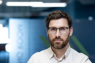 Gözlüklü ve sakallı genç bir adamın yakın plan portresi. Ofiste oturmuş, ciddi ve kendinden emin bir şekilde kameraya bakıyor..