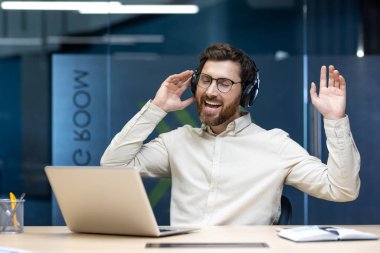Kulaklıklı rahat bir genç adam ofiste dizüstü bilgisayarla oturur ve dans ederken müzik dinler..