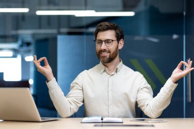 Ofiste çalışan rahat bir genç adam, mola sırasında lotus pozisyonunda masasına oturur ve meditasyon yapar, dizüstü bilgisayarın ekranına bakar..