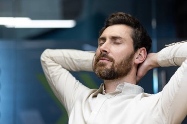 İş yerindeki sandalyede oturan genç bir ofis çalışanının yakın plan fotoğrafı. Elleri başının arkasında, gözleri kapalı ve dinleniyor..