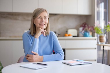 Üst düzey bir kadın mutfak masasında oturuyor, neşeli bir şekilde gülümserken, düşünceli bir şekilde başka tarafa bakıyor, rahat bir evde fikirleri ya da anıları yansıtıyor.