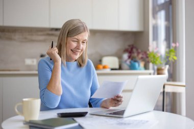 Mutfak masasında gülümseyen üst düzey bir kadın, faturaları ve belgeleri incelerken aynı zamanda bir dizüstü bilgisayarla bütçe oluşturur, tasarrufları yönetir ve güvenli, kendinden emin bir emeklilik geleceği planlar.