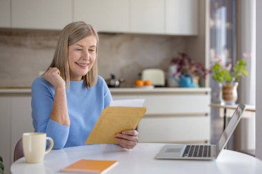 Üst düzey bir kadın heyecan verici bir belge ya da mektup okurken mutlu bir şekilde gülümsüyor, elinde dizüstü bilgisayarla bir masada oturuyor, evdeki neşe ve olumlu duyguları yansıtıyor.