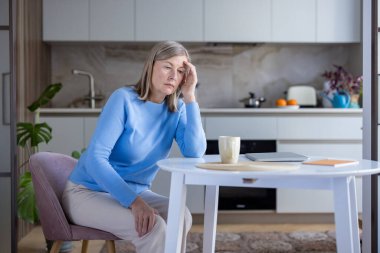 Mutfak masasında oturan olgun bir kadın stresli bir baş ağrısı yaşıyor, depresyonunu düşünürken yorgun ve yalnız hissediyor ve evinde huzur arıyor.
