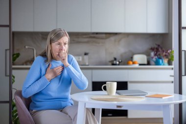 Yaşlı kadın kendini iyi hissetmiyor ve bir mutfak masasında otururken göğsünde bir elle acı verici bir öksürük yaşıyor. Solunum sorunları ya da hastalık belirtileri gösteriyor.