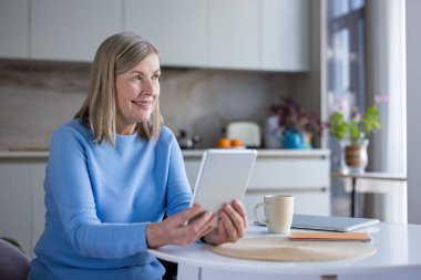 Açık mavi kazaklı yaşlı bir kadın mutfak masasına oturmuş gülümsüyor, elinde tabletle kahvenin tadını çıkarıyor ve sabah güneşinin aydınlığında internette geziniyor.