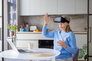 VR kulaklık takan üst düzey bir kadın sanal elementlerle etkileşime geçiyor ve ev mutfağındaki metaevreleri keşfediyor, modern teknolojinin ve gelecekteki eğlence dünyasının tadını çıkarıyor.