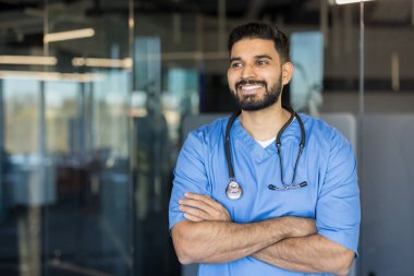 Mavi önlük giyen kendine güvenen genç erkek doktor ya da hemşire ve kolları bağlı steteskop, başka tarafa bakarken gülümseyen, kendini adamış sağlık uzmanlarını temsil eden