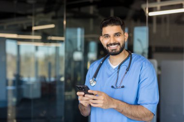 Mavi önlük ve steteskop giymiş gülümseyen erkek doktor, modern bir klinikte veya hastane ortamında kameraya neşeyle bakıyor ve akıllı telefon kullanıyor.