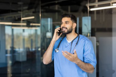Önlüklü ve steteskoplu erkek doktor cep telefonuyla konuşuyor, tele sağlık danışmanlığı yaparken veya modern ofis ortamında hasta bakımını tartışırken aktif iletişim kuruyor.