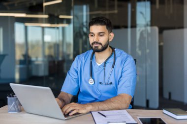 Tıp önlüğü ve steteskop giymiş genç Hintli erkek doktor bir masada dizüstü bilgisayar üzerinde çalışıyor, sağlık teknolojisi, online danışmanlık ve profesyonel bir klinikte teletıp hizmeti sunuyor.