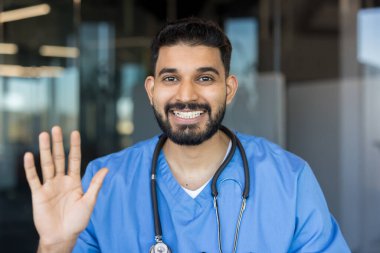 Dost canlısı genç bir erkek doktor ya da hemşire tıbbi önlük ve steteskop takmış gülümseyen ve el sallayan modern bir klinikte ya da hastanede duruyor.