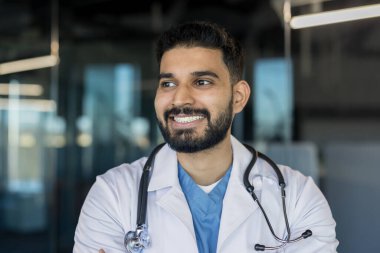 Steteskoplu beyaz önlüklü Hintli erkek doktor, sıcak bir şekilde gülümsüyor ve başka tarafa bakıyor, modern bir klinikte, profesyonel ve ulaşılabilir bir portrede duruyor.