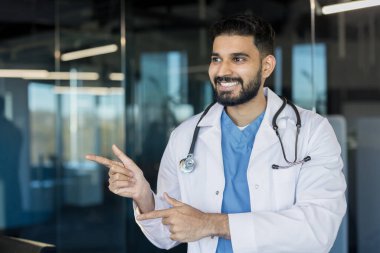 Steteskoplu beyaz önlüklü genç Hintli erkek doktor, özgüvenle gülümsüyor ve modern bir kliniği işaret ediyor, profesyonel rehberlik, bakım ve güvenilir sağlık hizmetlerini öneriyor.