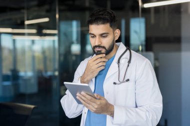 Beyaz önlüklü ve steteskoplu genç bir erkek doktor. Modern bir klinikte veya ofis ortamında dijital tabletin tıbbi bilgilerini inceliyorum. Yenilik ve profesyonel analizi temsil ediyor