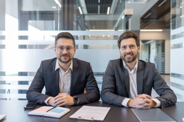 İki profesyonel iş ortağı bir konferans masasında oturuyor, modern ofis ortamında kurumsal bir toplantı sırasında kameraya gülümsüyor, işbirliği ve başarıyı temsil ediyor.