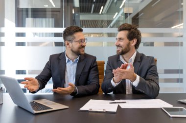 Bir konferans masasında gülümseyen iki işadamı, biri bir jestle konuşurken, diğeri başarılı takım çalışması, profesyonel işbirliği ve kutlamaları alkışlıyor
