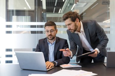 İki profesyonel iş adamı bir dizüstü bilgisayara bakıp ekranı göstererek, raporları inceleyerek ve modern bir ofiste yapılan bir şirket toplantısında yeni stratejileri tartışarak iş birliği yapıyorlar.