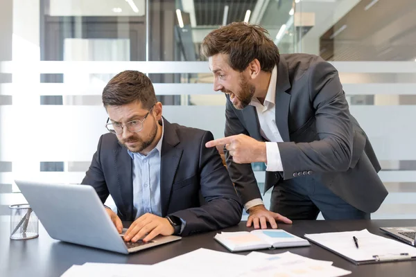 Öfkeli patron gergin bir ofis sahnesinde dizüstü bilgisayarla sinirli erkek çalışanı işaret ederek ve bağırarak işyerinde zorbalık, stres, baskı, çatışma ve ayrımcılık tasvir ediyor.