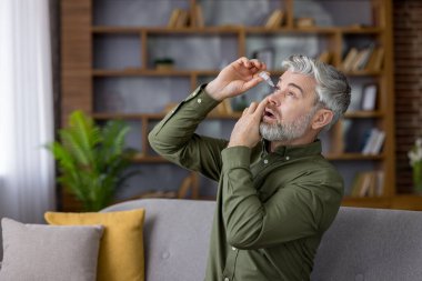 Olgun bir adam dikkatli bir şekilde evine göz damlası damlatarak, kuru gözlere ya da alerjik tepkilere dikkat ederek, kişisel sağlık, tedavi ve günlük rutine odaklanır.